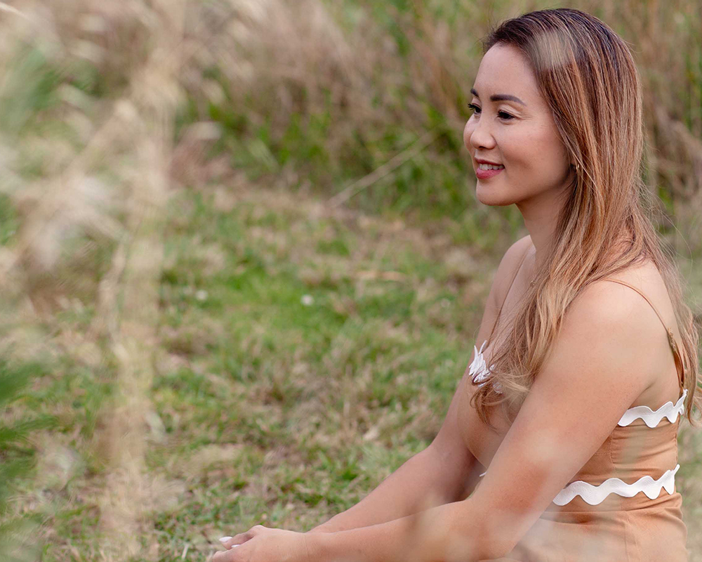 Portrait photo of Rasamy the celebrant sitting in a tan dress, from a profile view in a parkland setting and blurred grass in the foreground