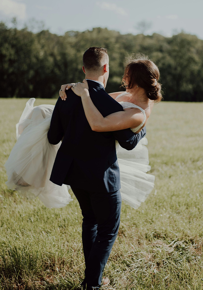 A groom carries a bride across a field from behind with a bank of trees in the distance