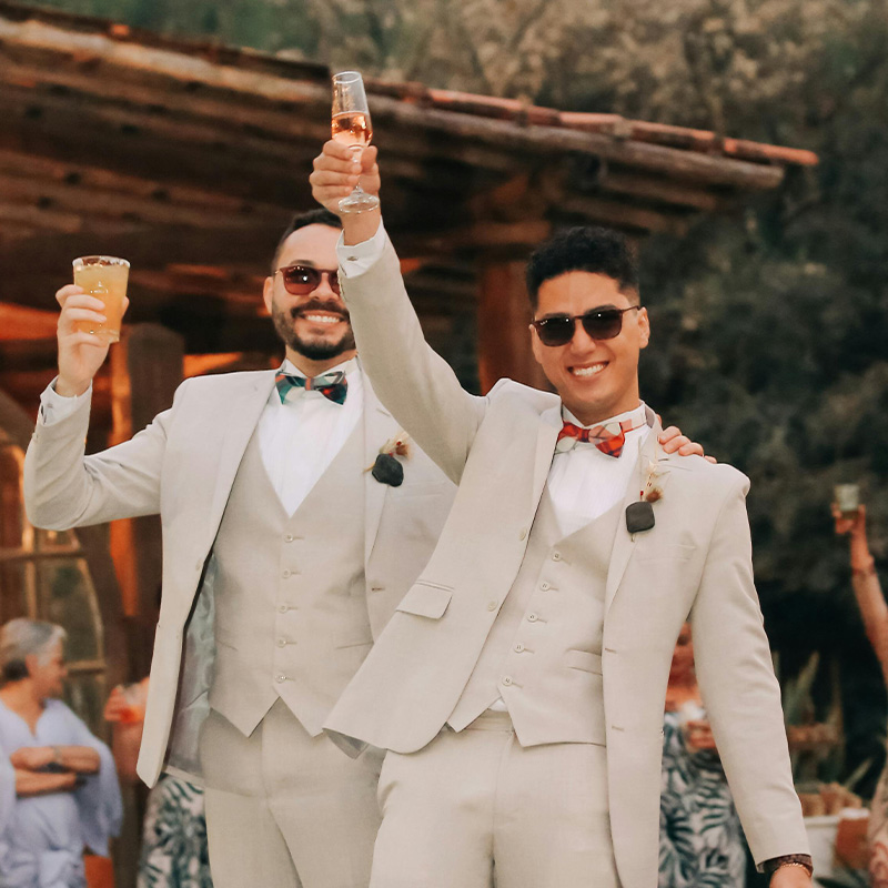 Two grooms in white suits and bow ties hold up champagne glasses happily after their wedding